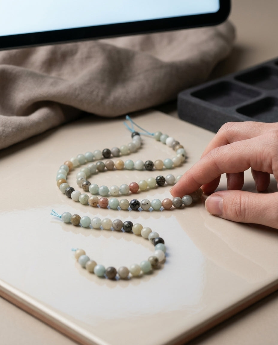 Beaded necklace on a table with a hand touching it, next to a computer screen.